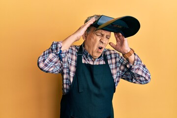 Handsome senior man with grey hair wearing welding protection mask suffering from headache desperate and stressed because pain and migraine. hands on head.