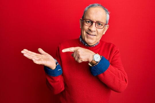 Handsome senior man with grey hair wearing casual clothes and glasses amazed and smiling to the camera while presenting with hand and pointing with finger.