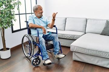 Handsome senior man sitting on wheelchair at the living room smiling with happy face looking and pointing to the side with thumb up.