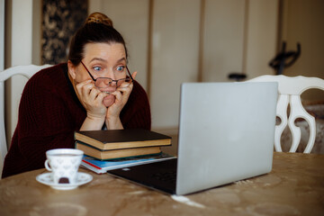 Portrait of frightened scared middle-aged woman sitting at table, looking at laptop, biting nails, watching horror film.