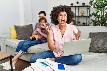 Mother of interracial family working using computer laptop at home crazy and mad shouting and yelling with aggressive expression and arms raised. frustration concept.