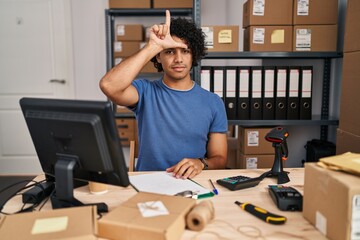 Hispanic man with curly hair working at small business ecommerce making fun of people with fingers on forehead doing loser gesture mocking and insulting.