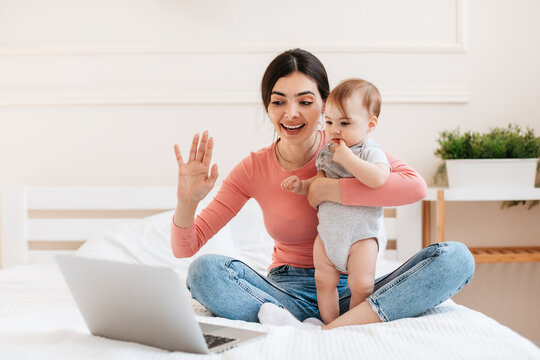 Happy Mom Holding Baby And Making Video Call With Laptop, Sitting On Bed At Home And Waving Hand At Webcamera