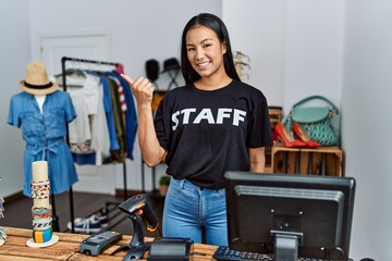 Young hispanic woman working as staff at retail boutique pointing to the back behind with hand and thumbs up, smiling confident