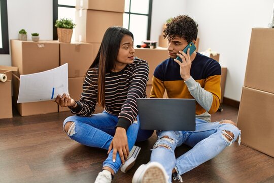 Young Latin Couple Using Laptop And Talking On The Smartphone Sitting On The Floor At New Home.