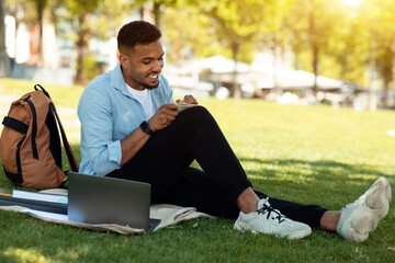 Happy black male student studying remotely with laptop and writing down information in notebook, sitting in park © Prostock-studio