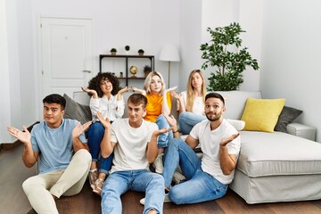 Group of people sitting on the sofa and floor at home clueless and confused expression with arms...