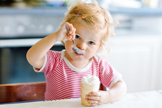 Adorable baby girl eating from spoon sweet ice cream in waffle cone. food, child, feeding and development concept. Cute toddler, daughter with spoon sitting in highchair and learning eat by itself