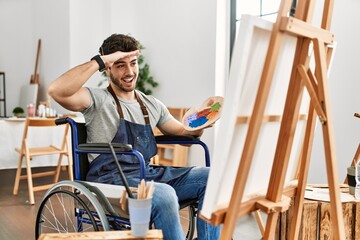 Fototapeta na wymiar Young hispanic man sitting on wheelchair painting at art studio very happy and smiling looking far away with hand over head. searching concept.