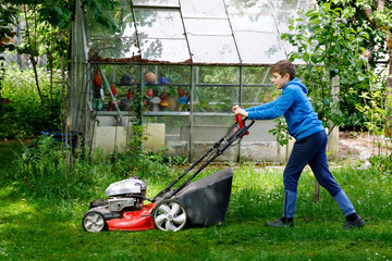 Happy preteen kid boy with lawn mower. Portrait of smiling teenager child working in garden,...