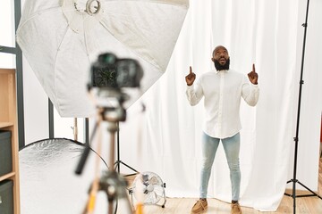 African american man posing as model at photography studio amazed and surprised looking up and pointing with fingers and raised arms.