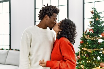Young interracial couple kissing and hugging standing by christmas tree at home.