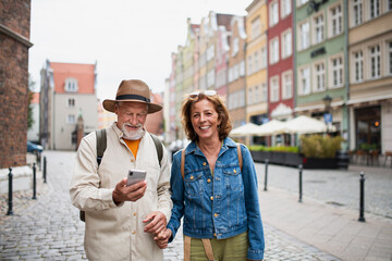 Portrait of happy senior couple tourists smiling, holding hands, using smartphone outdoors in historic town