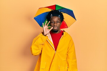 Young african american man wearing yellow raincoat mouth and lips shut as zip with fingers. secret...