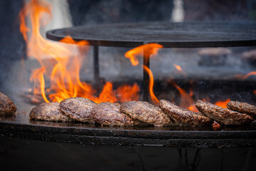 Bunch of patties flattened round, serving of minced ground beef meat being prepared on outdoor barbeque grill with bright orange flame at kitchen of street food restaurant used for burgers cooking
