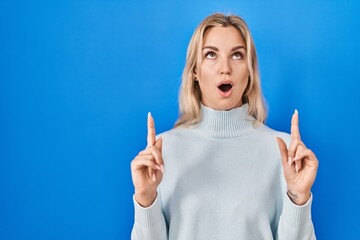 Young caucasian woman standing over blue background amazed and surprised looking up and pointing with fingers and raised arms.