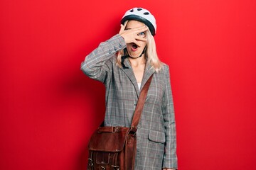 Beautiful caucasian blonde business woman wearing bike helmet peeking in shock covering face and...