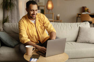 Joyful young black man with passport and ticket using laptop, booking abroad vacation at online tourist agency from home