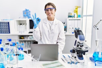 Young hispanic woman wearing scientist uniform working at laboratory doing ok sign with fingers, smiling friendly gesturing excellent symbol - obrazy, fototapety, plakaty