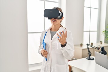 Young blonde woman wearing doctor uniform using touchpad and vr goggles at clinic