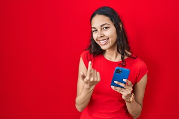 Young brazilian woman using smartphone over red background beckoning come here gesture with hand inviting welcoming happy and smiling