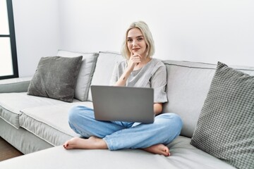 Young caucasian woman using laptop at home sitting on the sofa looking confident at the camera smiling with crossed arms and hand raised on chin. thinking positive.