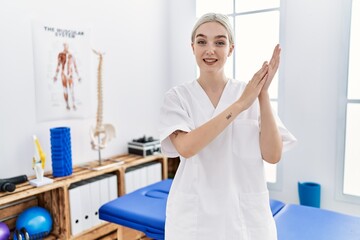 Young caucasian woman working at pain recovery clinic clapping and applauding happy and joyful, smiling proud hands together