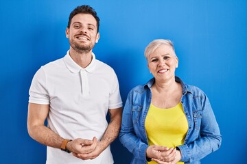 Young brazilian mother and son standing over blue background with hands together and crossed fingers smiling relaxed and cheerful. success and optimistic