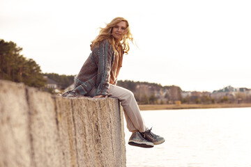 Portrait of young smiling woman with long wavy fair hair sitting with crossed legs on edge of concrete river embankment.