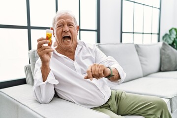 Senior man holding pills angry and mad screaming frustrated and furious, shouting with anger. rage...