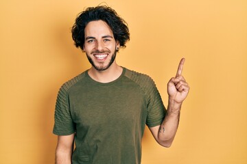 Handsome hispanic man wearing casual green t shirt smiling with an idea or question pointing finger up with happy face, number one