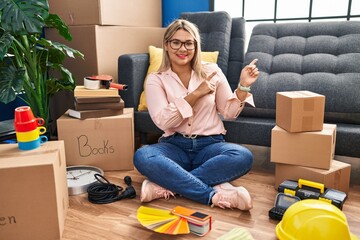 Young hispanic woman moving to a new home sitting on the floor smiling and looking at the camera pointing with two hands and fingers to the side.