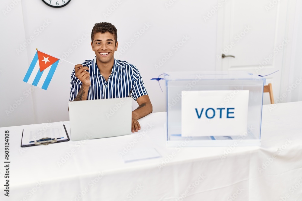 Canvas Prints Young handsome hispanic man at political campaign election holding cuba flag looking positive and happy standing and smiling with a confident smile showing teeth