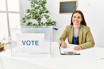 Young brunette woman sitting at election table with voting ballot with a happy and cool smile on...