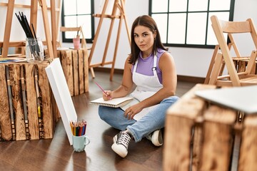 Young beautiful hispanic woman at art studio thinking attitude and sober expression looking self confident