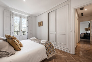 Double bedroom with beautiful decoration in hardwood, white fitted wardrobes, matching blanket and cushions, herringbone oak flooring and balcony with white wooden windows and shutters.