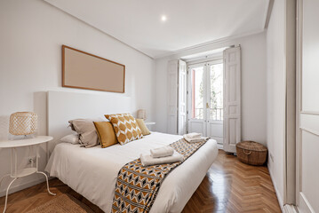 Double bedroom with bed with white duvet, matching blanket and cushions, white with wooden windows and herringbone oak flooring