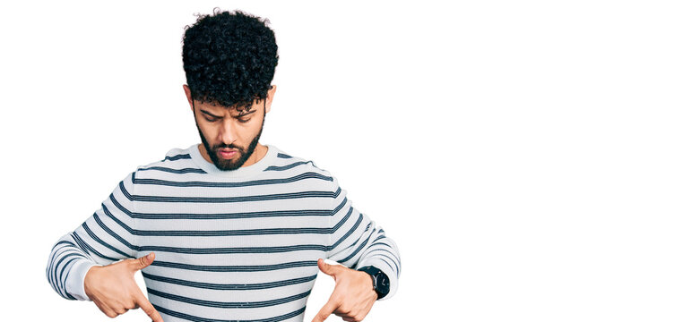 Young arab man with beard wearing casual striped sweater pointing down with fingers showing advertisement, surprised face and open mouth
