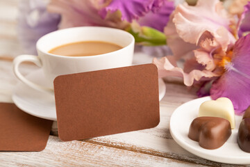 Brown business card with cup of cioffee, chocolate candies and iris flowers on white wooden background. side view, copy space.