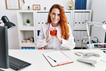 Young redhead woman wearing doctor uniform holding heart at hospital