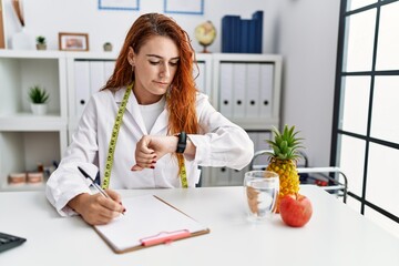 Young redhead woman nutritionist doctor at the clinic checking the time on wrist watch, relaxed and...