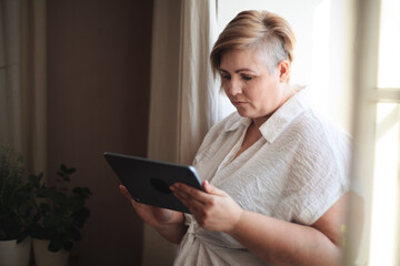 Overweight woman in white shirt standing by window and using tablet.
