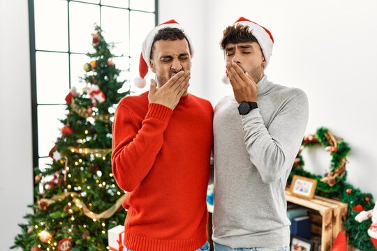 Young gay couple standing by christmas tree wearing hat bored yawning tired covering mouth with hand. restless and sleepiness.