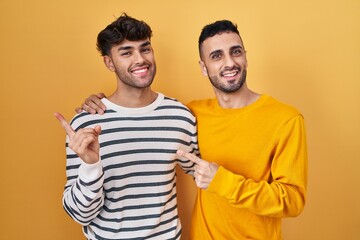 Young hispanic gay couple standing over yellow background cheerful with a smile on face pointing with hand and finger up to the side with happy and natural expression