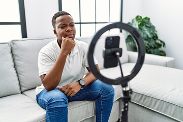 Young african man recording vlog tutorial with smartphone at home serious face thinking about...