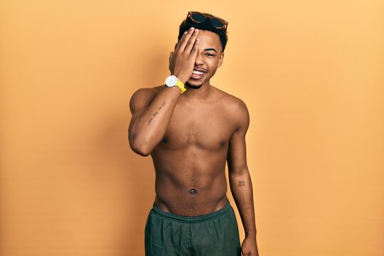 Young african american man wearing swimwear and swimmer glasses covering one eye with hand, confident smile on face and surprise emotion.