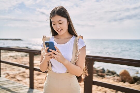 Young chinese girl smiling happy using smartphone at the beach.