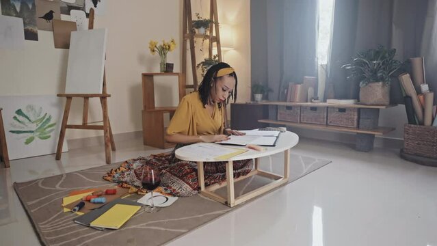 Young black woman drawing picture at table while sitting on floor with wine glass and craft tools in living room