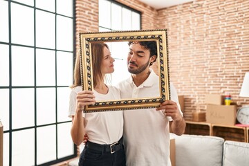 Young two people holding empty frame together smiling looking to the side and staring away thinking.