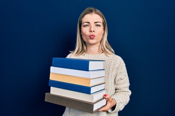 Beautiful caucasian woman holding a pile of books making fish face with mouth and squinting eyes, crazy and comical.